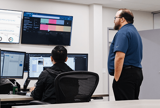 Two IT professionals monitor system dashboards across several screens in a bright office setting.